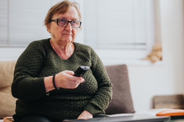 An elderly woman wearing glasses sits comfortably on a couch holding a remote control, watching TV....