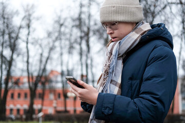 Teenager Girl Walking Alone on City Street with Smartphone. Portrait of 14 years old girl wearing eyeglasses, wool hat, winter jacket using mobile phone. Urban Style. Rainy mood weather. Cold season