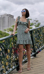 Stylish Asian woman wearing a floral dress and high-heeled sandals stands on a decorative city bridge with ornate railings and lush greenery