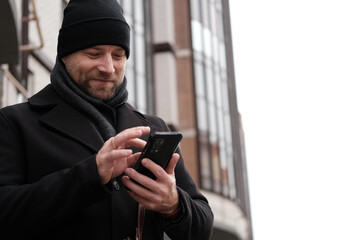 Handsome man wearing black coat and brown leather bag walking down on the city with mobile phone in hand. Stylish bearded middle-aged male using smartphone at urban street. Businessman going on job.