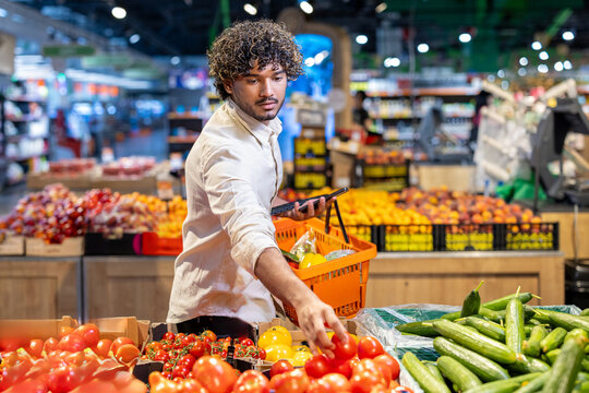 A man with curly hair shops for fresh produce, including tomatoes and cucumbers, in a well-lit grocery store.