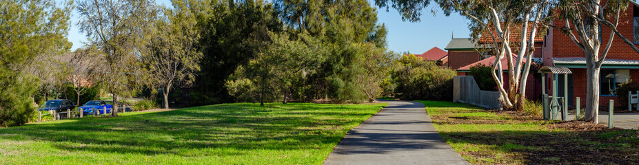 Panoramic view of a quiet walking path runs alongside a well-maintained lawn and mature trees in a suburban residential neighborhood.  A public green space in Melbourne Australia's local neighborhood.