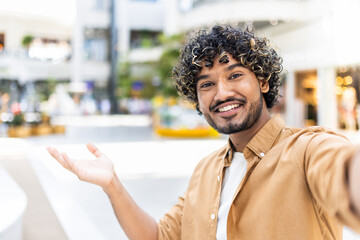 A smiling man with curly hair takes a selfie outside, extending his arm towards the viewer,...