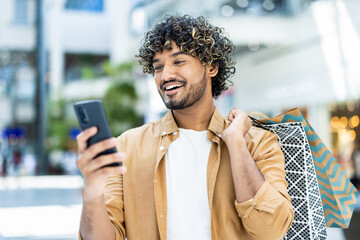 A young man looks at his phone while shopping in a mall, holding shopping bags over his shoulder,...