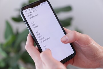 Cyber bullying. Woman holding smartphone with insults on blurred background, closeup