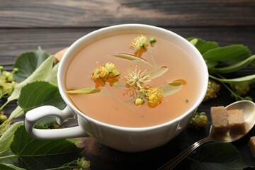 Fresh linden tea in cup, sugar cubes and flowers on dark wooden table, closeup