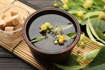Fresh linden tea in cup, sugar cubes and flowers on dark wooden table, closeup