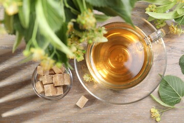 Tasty linden tea in cup, sugar cubes, leaves and flowers on wooden table, flat lay
