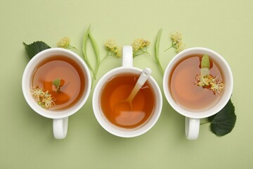 Tasty linden tea in cups, spoon, leaves and flowers on light green background, flat lay
