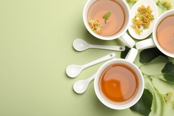 Tasty linden tea in cups, spoons, leaves and flowers on light green background, flat lay. Space for...