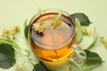 Tasty linden tea in cup, leaves and flowers on light green background, closeup