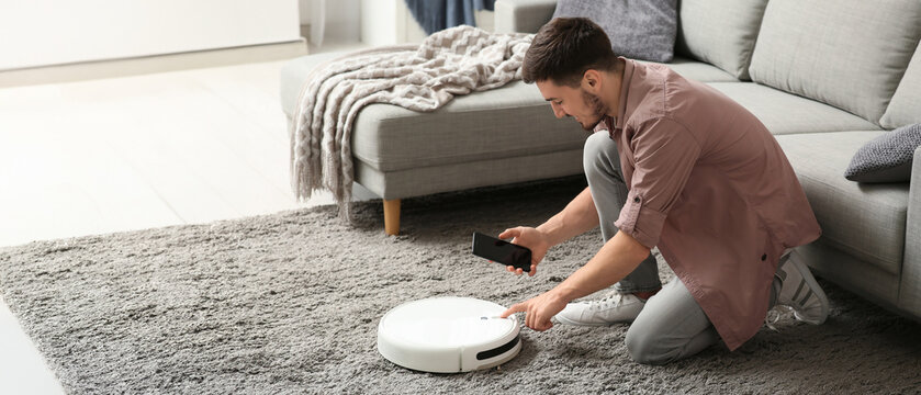 Young man with mobile phone turning on modern robot vacuum cleaner at home - Powered by Adobe