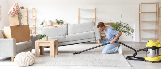 Male janitor cleaning floor under sofa with vacuum cleaner in room