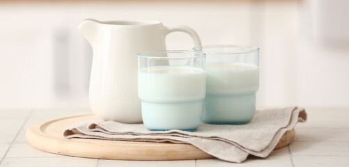 Wooden board with ceramic jug and glasses of milk on white tile table