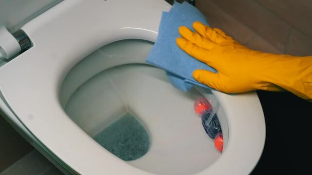 A man wearing yellow rubber work gloves washes the toilet and toilet seat with a blue antibacterial cloth to kill harmful germs and bacteria.
Cleaning and disinfecting the toilet