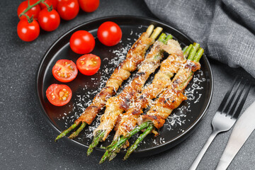 Grilled asparagus wrapped in crispy bacon sprinkled with parmesan cheese served on plate with fresh cherry tomatoes on black background. Gourmet dish, cooking recipe, cuisine, or restaurant menu