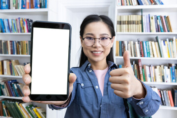 An asian woman is holding a blank phone screen in front of a bookshelf, giving a thumbs up and...