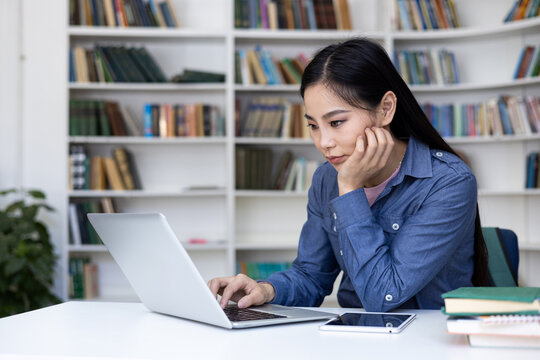 A focused Asian woman works on a laptop in a library, surrounded by books and a digital tablet.