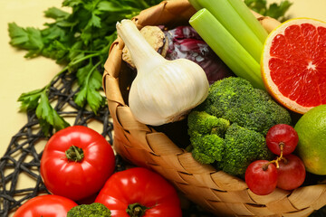 Wicker basket with different fresh fruits and vegetables on yellow background, closeup
