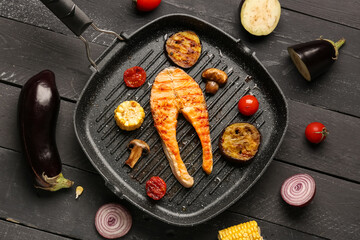 Frying pan with tasty grilled salmon steak and vegetables on black wooden background