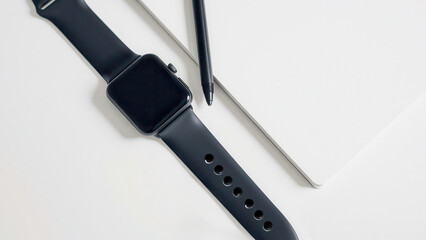 Black smartwatch and stylus lie next to a notepad on a white table. Modern business style. The theme of productivity, planning and digital gadgets.