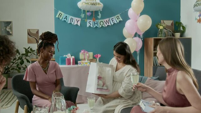 Full shot of group of multiethnic girlfriends sitting around table with sweets at baby shower party. African American guest giving pregnant Hispanic woman baby hamper, admiring tiny pair of shoes