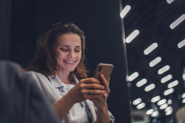 Smiling young adult woman using mobile phone in building with night lighting