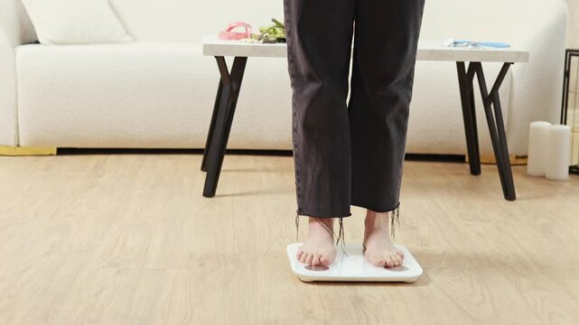 Woman stepping on body composition scale in living room with sofa and table. Girl checking weight, making effort to lose kilos. Slim and fit body goal, diet and health concept.