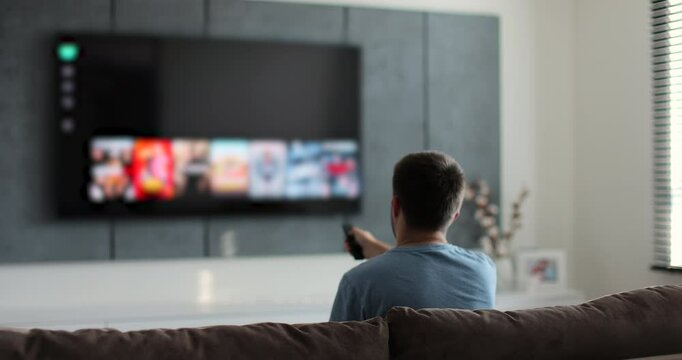 Man selects tv channels with remote control at living room on sofa. Young man controls TV using a modern remote control.
