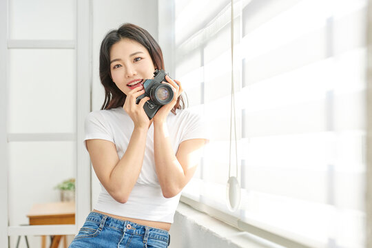 Young Asian Woman Photographer in White T-Shirt Holding Vintage Camera by Bright Window Indoors