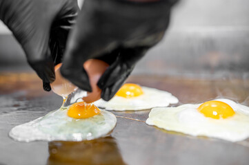 Close-up of a person in black gloves cracking an egg onto a hot griddle with two eggs already cooking, capturing the moment of preparation in a professional kitchen setting