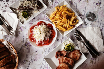 Traditional Balkan meal served on a rustic table with grilled meat, fries, tomato salad, onions, and fresh bread. Top view.