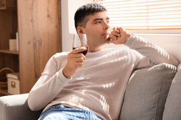 Thoughtful young man with glass of red wine sitting on sofa at home