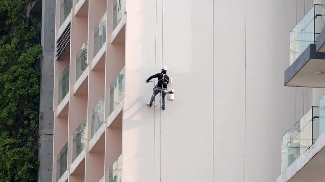 A professional worker with safety equipment paints the tall exterior wall of a modern high-rise building