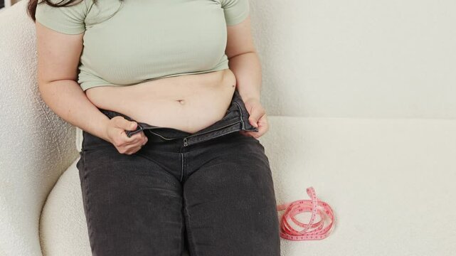 Woman holding belly fat with one hand and body composition scale in another, measuring tape on neck. Girl with unbuttoned pants wants to lose weight and get slim and fit.
