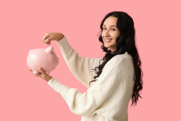 Pretty young woman putting coin into piggy bank on pink background