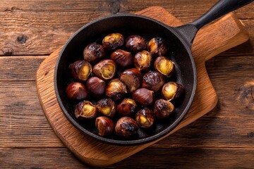 A cast iron pan filled with roasted chestnuts rests on a wooden board. The nuts showcase a crispy exterior and toasted aroma, perfect for cozy autumn gatherings