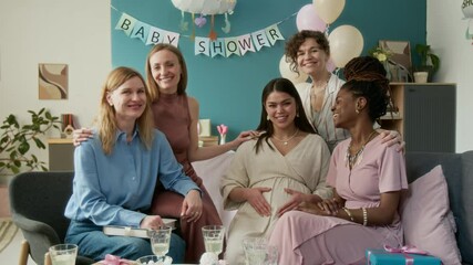 Medium full portrait of happy young heavily pregnant woman in beige dress sitting on couch at baby shower, caressing baby belly, surrounded by smiling diverse girlfriends, looking at camera