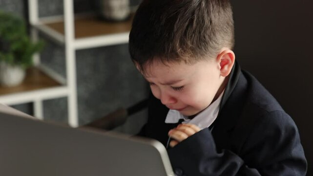 Baby boss sitting in office on chair working at computer and laptop. Kid playing businessman role, crying from tiredness, then relaxing on orange sofa in bright room with blinds and sunlight.