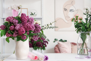 Fototapeta premium Fresh purple lilac flowers in white vase with eucalyptus branches and craft paper rolls on white table, the floral display creates a serene and inviting atmosphere in a modern interior setting