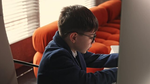 Adorable boy with glasses sitting on office chair pretending to work. School age kid in suit typing on keyboard in room with orange sofa, blinds, and sunlight.