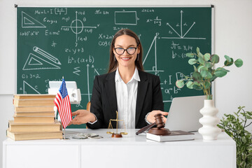 Female teacher with bribe, justice scales and judge's gavel on table in classroom. Corruption...