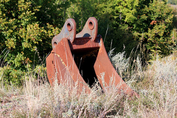 Large rusted excavator bucket stands upright among tall dry grasses and wild plants, abandoned at...