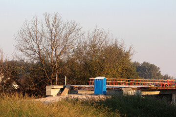 Bright blue portable toilet stands on a concrete slab at a rural construction site, surrounded by...