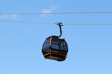 Contemporary gondola lift cabin with tinted windows and orange trim hangs from a cable, isolated against a clear blue sky, symbolizing modern transport and mountain tourism