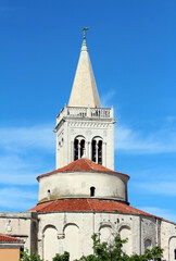 Fototapeta premium Tall stone church tower with pointed spire and arched windows rises above a round nave with red tiled roof, set against a clear blue sky in a historic European city
