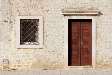 Historic stone building features a double wooden door and a square window with metal bars, framed...