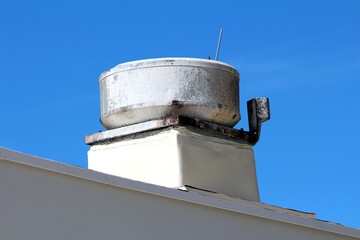 Large round metal chimney cap with rust and stains sits atop a flat white roof, mounted on a square...