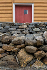 A weathered stone wall made of various rocks stands in front of an orange barn featuring a bold red door under overcast skies.