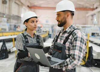 Two factory workers discussing production with laptop in a large industrial building